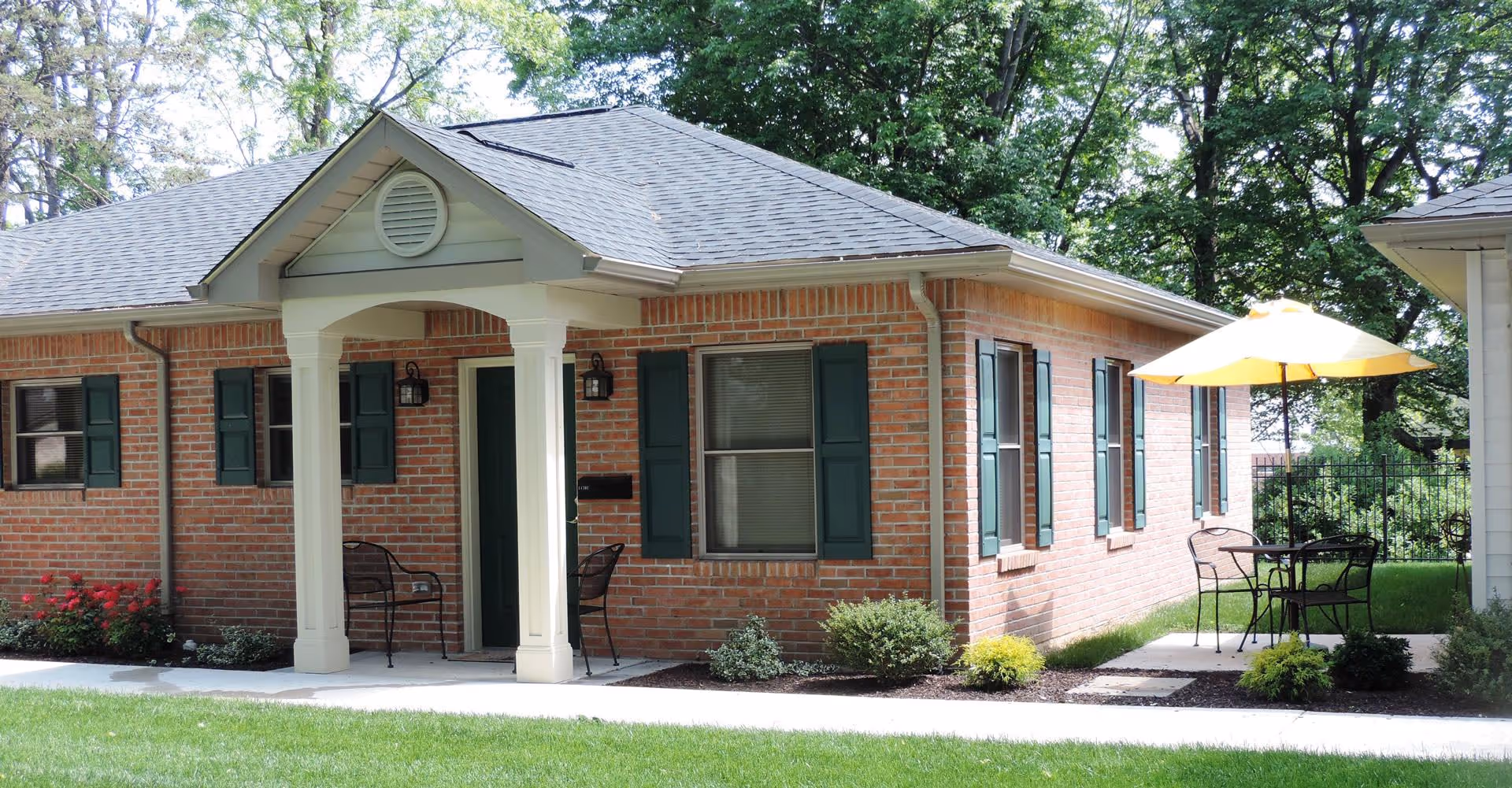 Exterior view of a single-story brick building with green shutters and a small covered porch with two chairs. There is a patio area with a table, chairs, and a yellow umbrella. The building is surrounded by green grass, bushes, and trees.