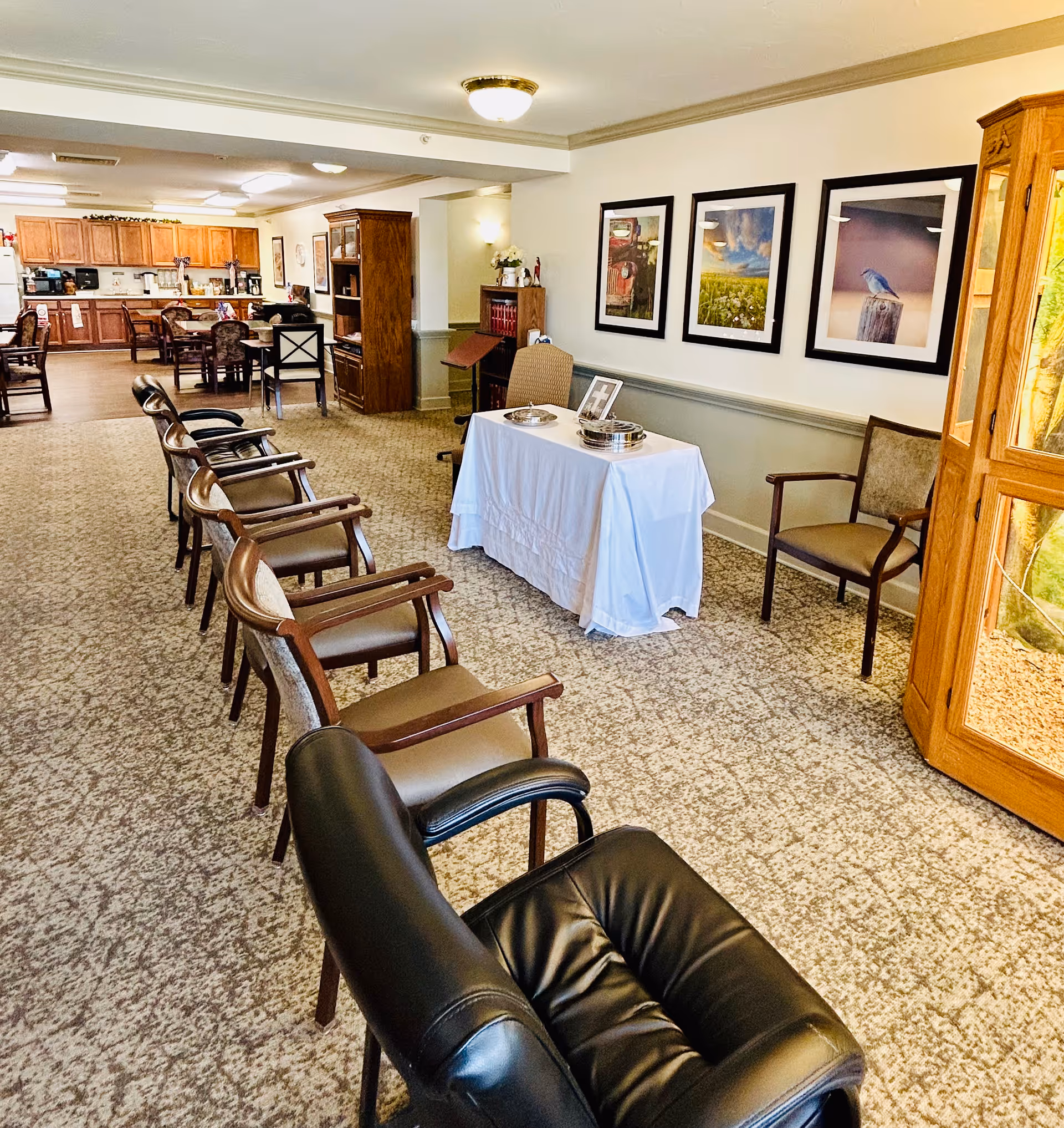 A communal senior living lounge with a row of chairs facing a small table draped in white, framed wall art, and a kitchenette/dining area in the background.