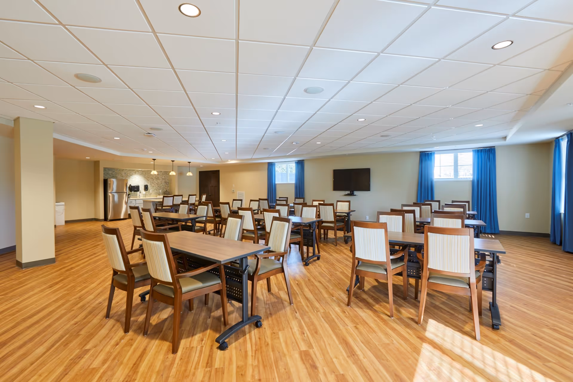 A spacious dining room with multiple wooden tables and chairs arranged neatly on a wood-patterned floor. The room has beige walls, a white tiled ceiling with recessed lighting, and two windows with blue curtains letting in natural light. There is a wall-mounted TV and a small kitchenette area with a refrigerator and pendant lights in the background.