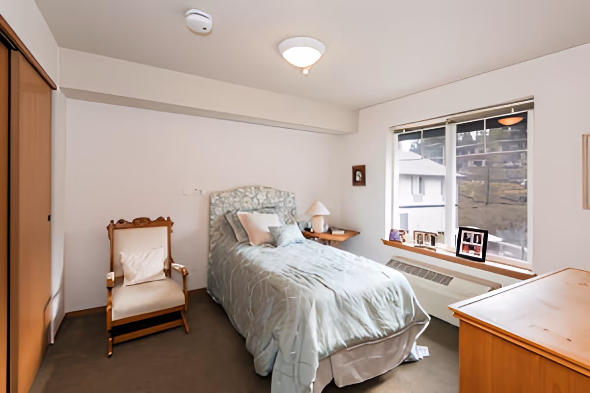 A small bedroom with a single bed covered in a light blue comforter and multiple pillows. Next to the bed is a wooden side table with a lamp. There is a wooden armchair with a cushion to the left of the bed. A large window with a view outside is on the right wall, with several framed photos on the windowsill. A wooden dresser is partially visible in the foreground on the right side. The room has white walls and a ceiling light fixture.