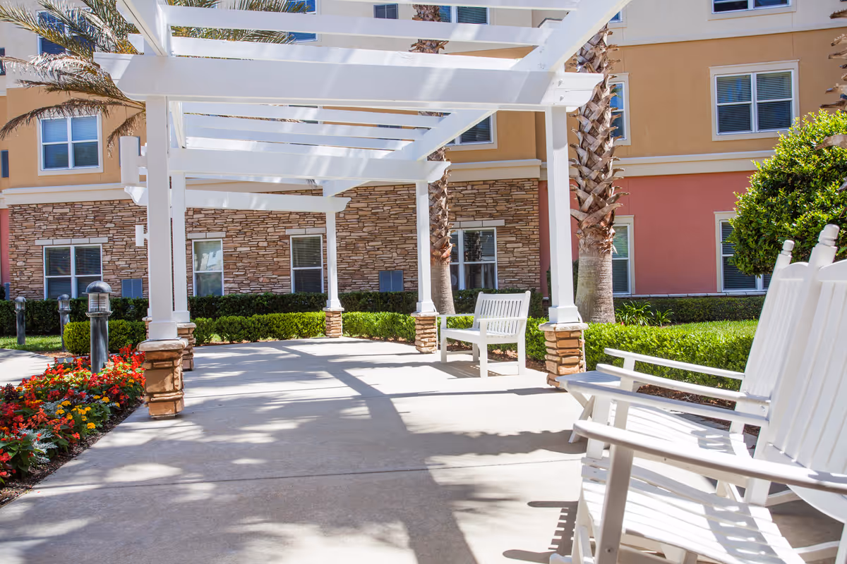 Outdoor seating area at Camellia At Deerwood featuring white wooden rocking chairs and benches under a white pergola, surrounded by palm trees, green bushes, colorful flowers, and a building with stone and stucco exterior walls and multiple windows.