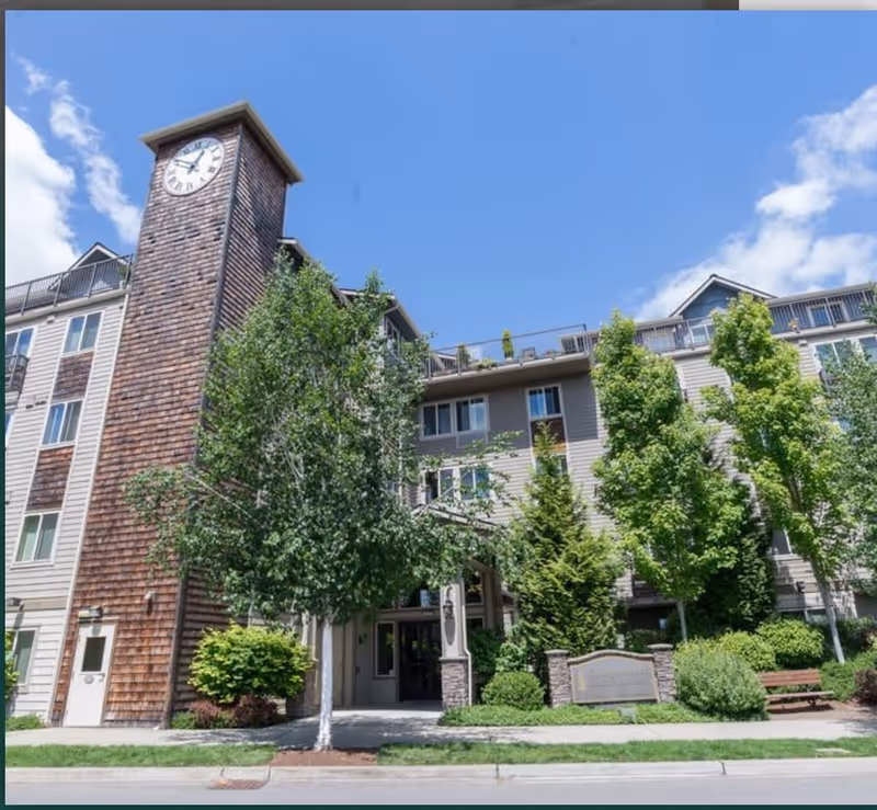 Exterior view of Covington Place Senior Apartments building with a prominent clock tower, surrounded by green trees and landscaping under a blue sky with some clouds.
