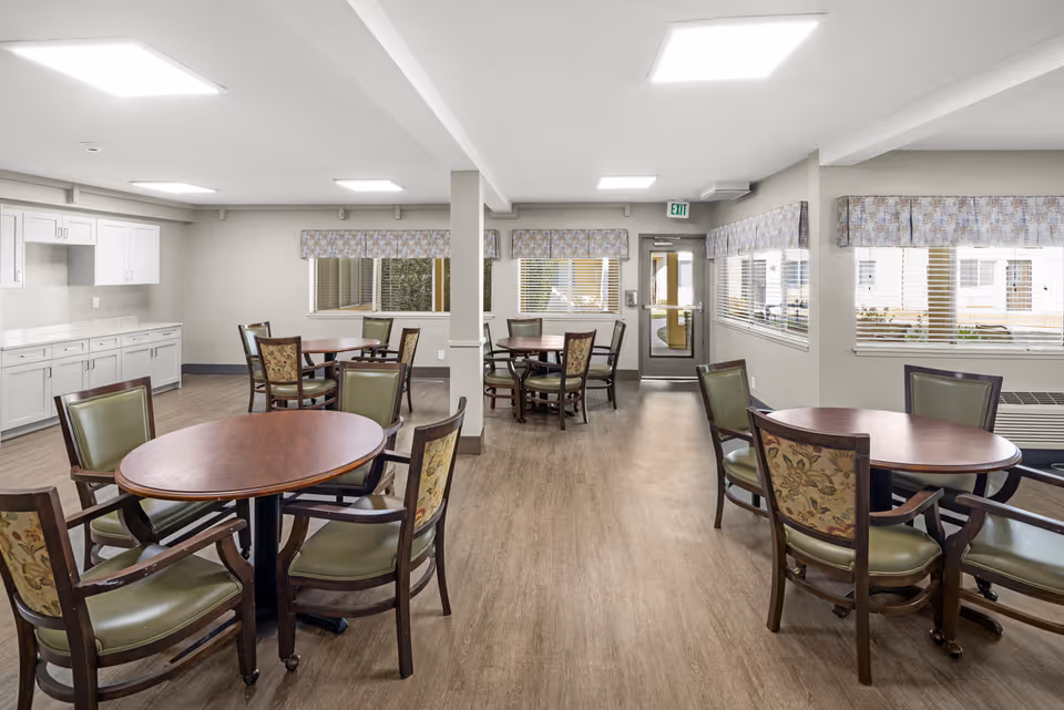 A bright and clean dining room in a senior living community with several round wooden tables surrounded by chairs with green and floral upholstery. The room has large windows with patterned valances, light wood flooring, white walls, and a white ceiling with recessed lighting. There is a door at the far end and white cabinetry along one wall.