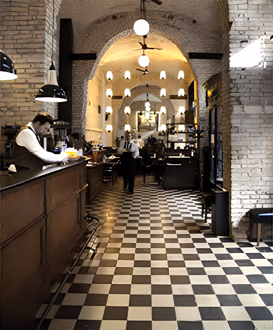 Arched interior café or dining space with a black-and-white checkered floor, a service counter on the left, and pendant lights hanging from the ceiling.