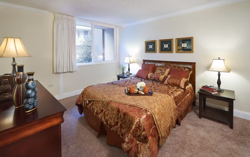 A cozy bedroom with a large bed covered in a patterned red and gold bedspread and multiple pillows. There are two bedside tables, each with a lamp, and a window with beige curtains letting in natural light. A dresser with decorative vases and a lamp is visible on the left side of the room.