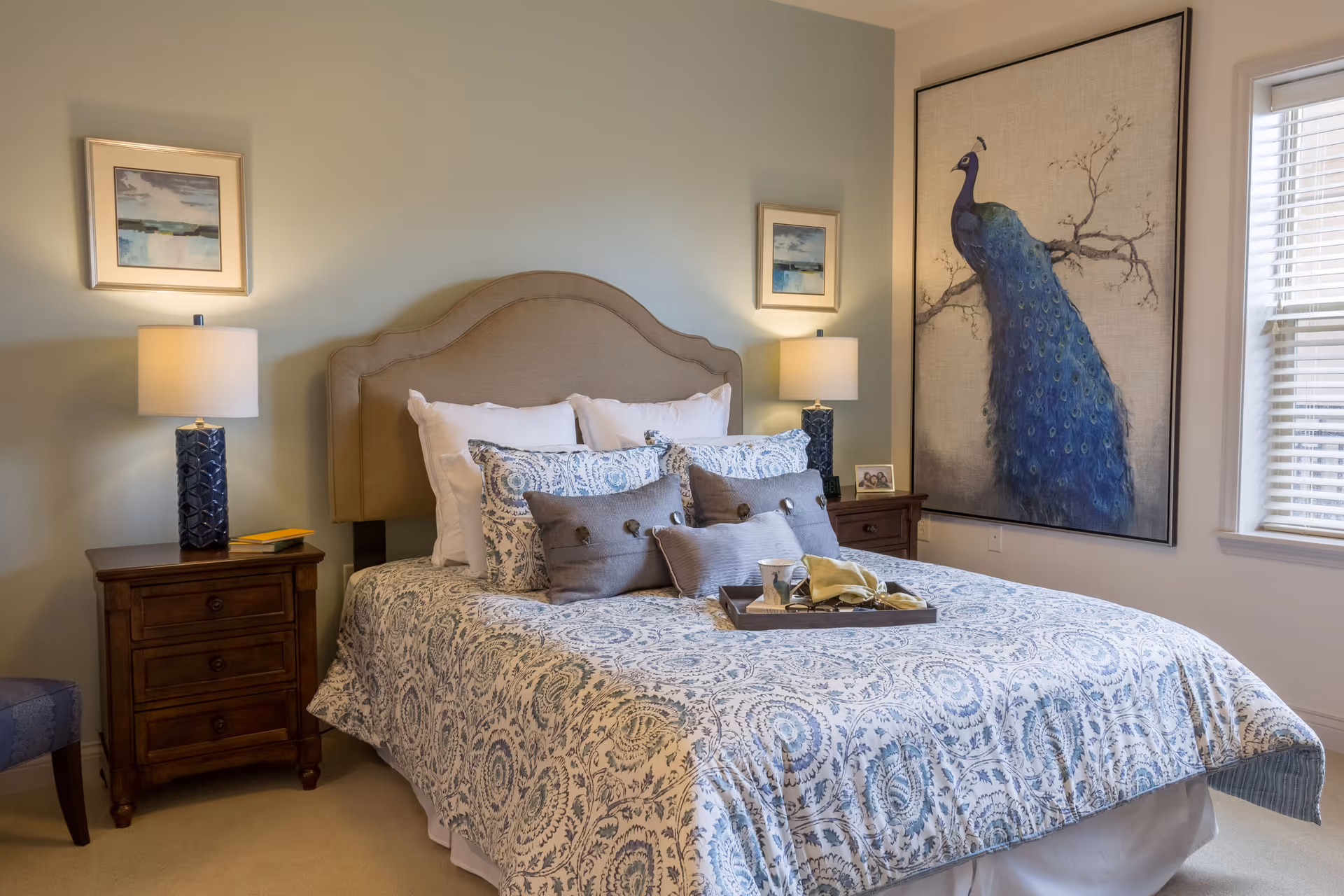 A neatly made bedroom with a patterned bed, upholstered headboard, bedside tables with lamps, and a large peacock artwork on the wall.
