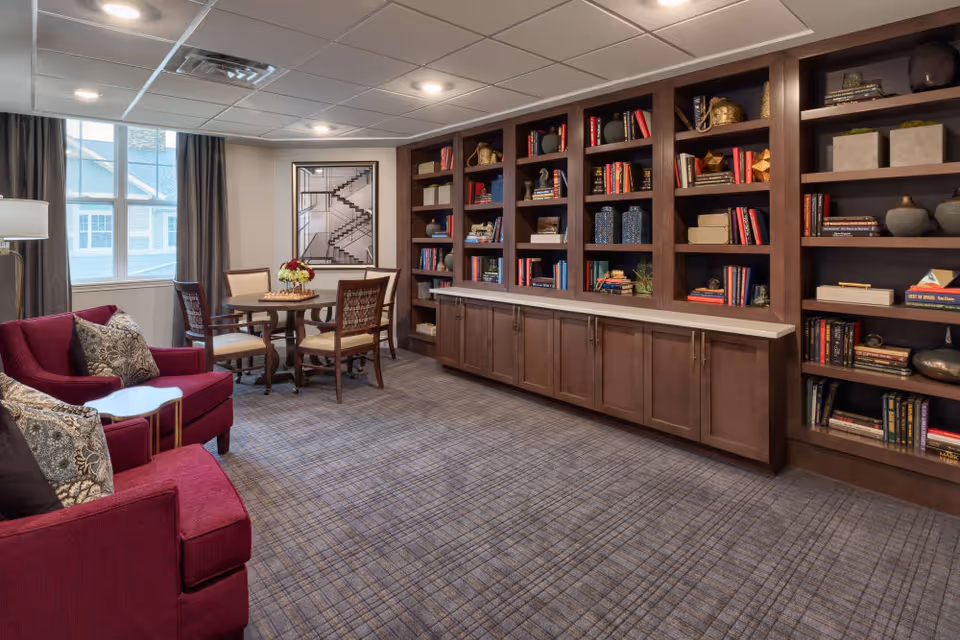 A cozy interior room featuring a large built-in wooden bookshelf filled with books and decorative items along one wall. In front of a window with curtains, there is a round wooden table with four chairs. Two red upholstered armchairs with patterned cushions are positioned near the table. The room has a carpeted floor with a plaid pattern and a ceiling with recessed lighting.