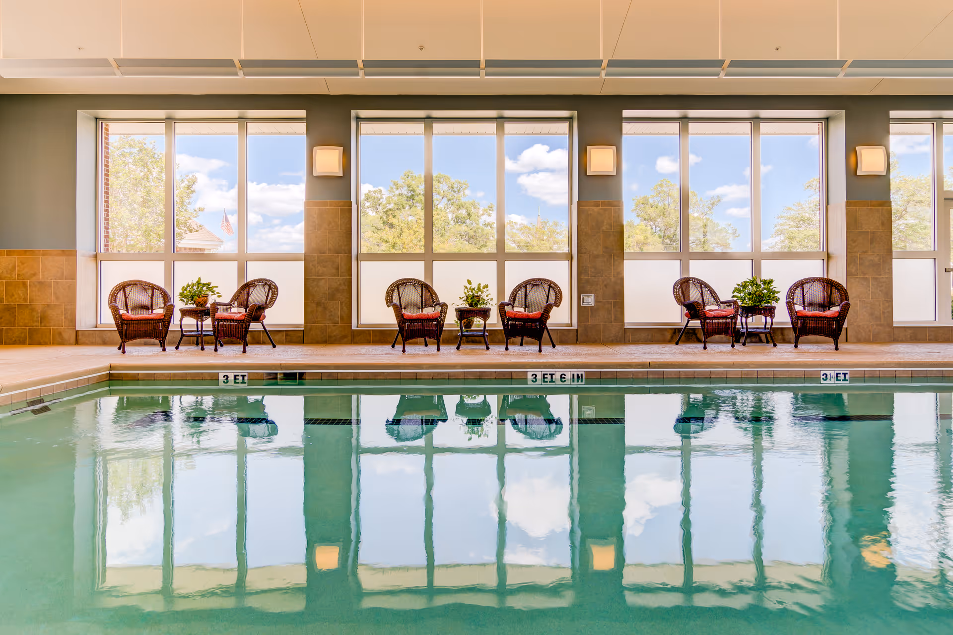 Indoor swimming pool with clear water reflecting large windows. Along the poolside, there are several sets of wicker chairs with red cushions and small tables, each adorned with green plants. The windows show a view of trees and a partly cloudy sky outside.