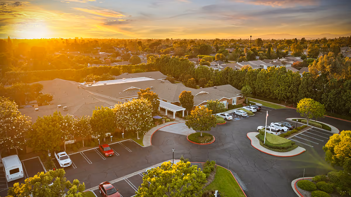 Aerial view of Ivy Park at Tustin senior living facility during sunset, showing the building surrounded by trees, a parking lot with several cars, and a landscaped circular driveway with an American flagpole.