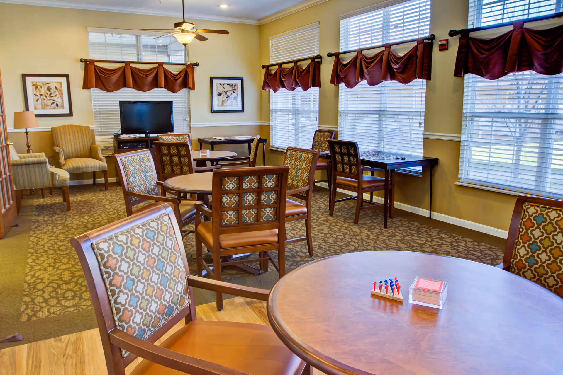 Community lounge with round tables and patterned chairs, a TV, and large windows with blinds.