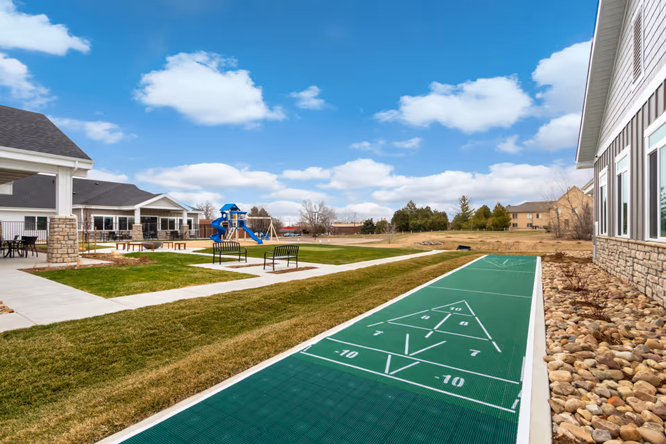 Outdoor recreational area at Greeley Village featuring a shuffleboard court in the foreground, green grass, benches, a playground with blue slides and swings, and buildings with stone and siding exteriors under a partly cloudy sky.