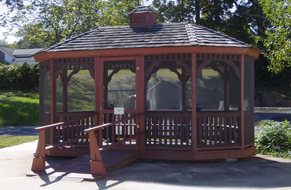 A wooden gazebo with a shingled roof and screened sides, situated outdoors on a concrete surface with greenery and trees in the background. The gazebo has a small ramp leading to its entrance.