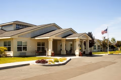 Exterior view of Settler's Park Senior Living facility showing a single-story building with a covered entrance, landscaped greenery, and an American flag on a flagpole under a clear blue sky.