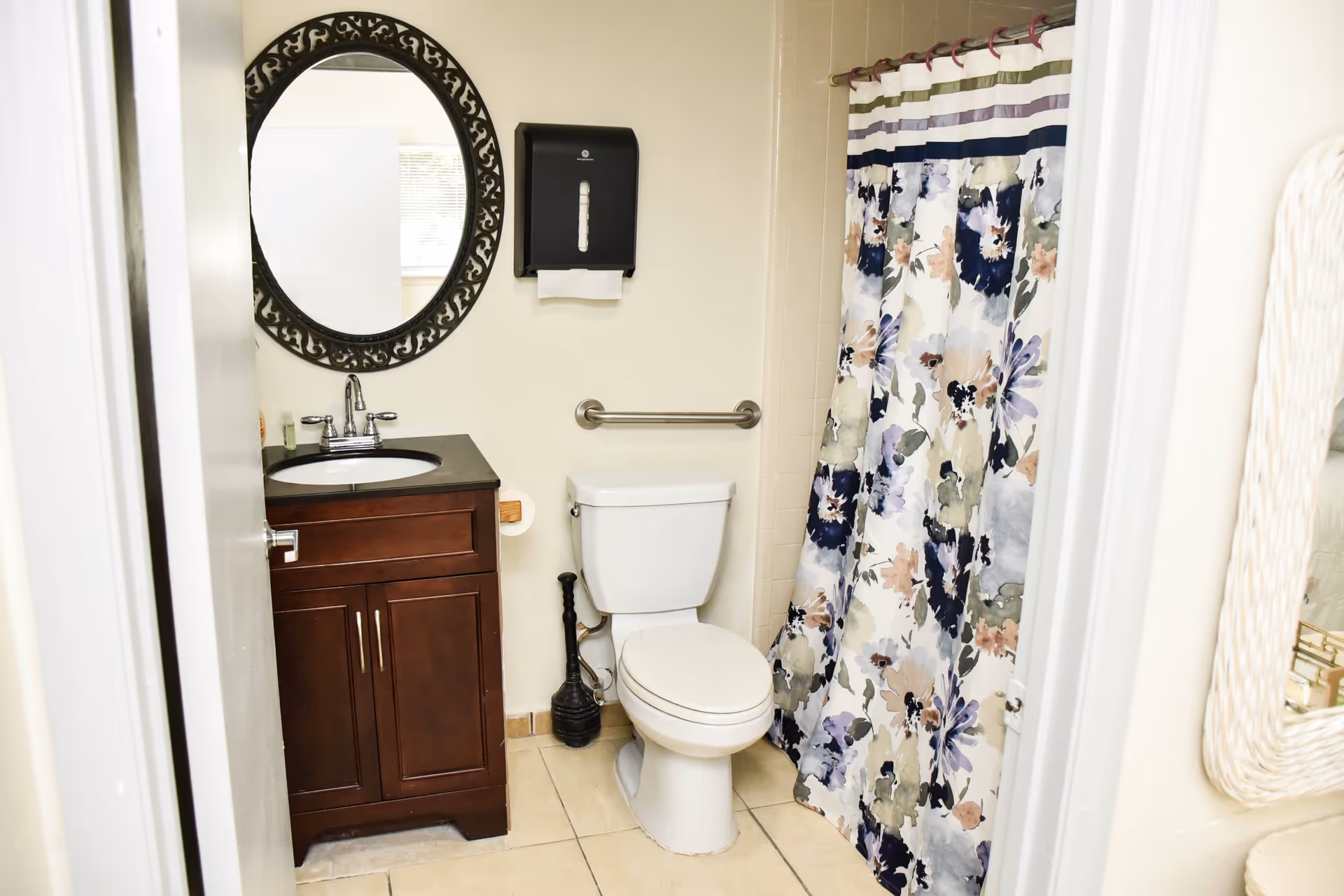 A small bathroom with a white toilet, a dark wood vanity with a black countertop and a round decorative mirror above it. There is a floral patterned shower curtain on the right side and a black paper towel dispenser mounted on the wall above the toilet. A grab bar is installed on the wall behind the toilet.