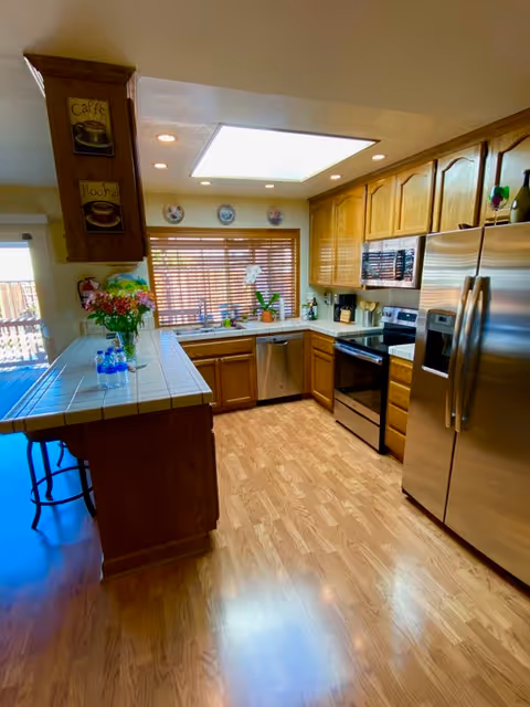 A bright kitchen with wooden cabinets, stainless steel refrigerator, oven, microwave, and dishwasher. The kitchen has a tiled countertop with flowers and water bottles on it, a large window with wooden blinds, and a wooden floor.