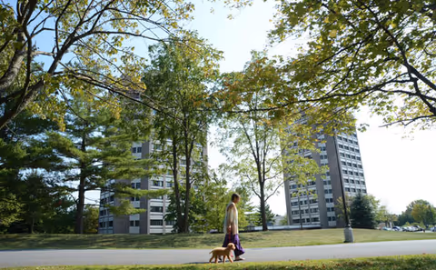 A person walking a small dog on a leash along a paved path in a park-like setting with green grass and trees. In the background, there are two tall apartment buildings partially obscured by the trees.