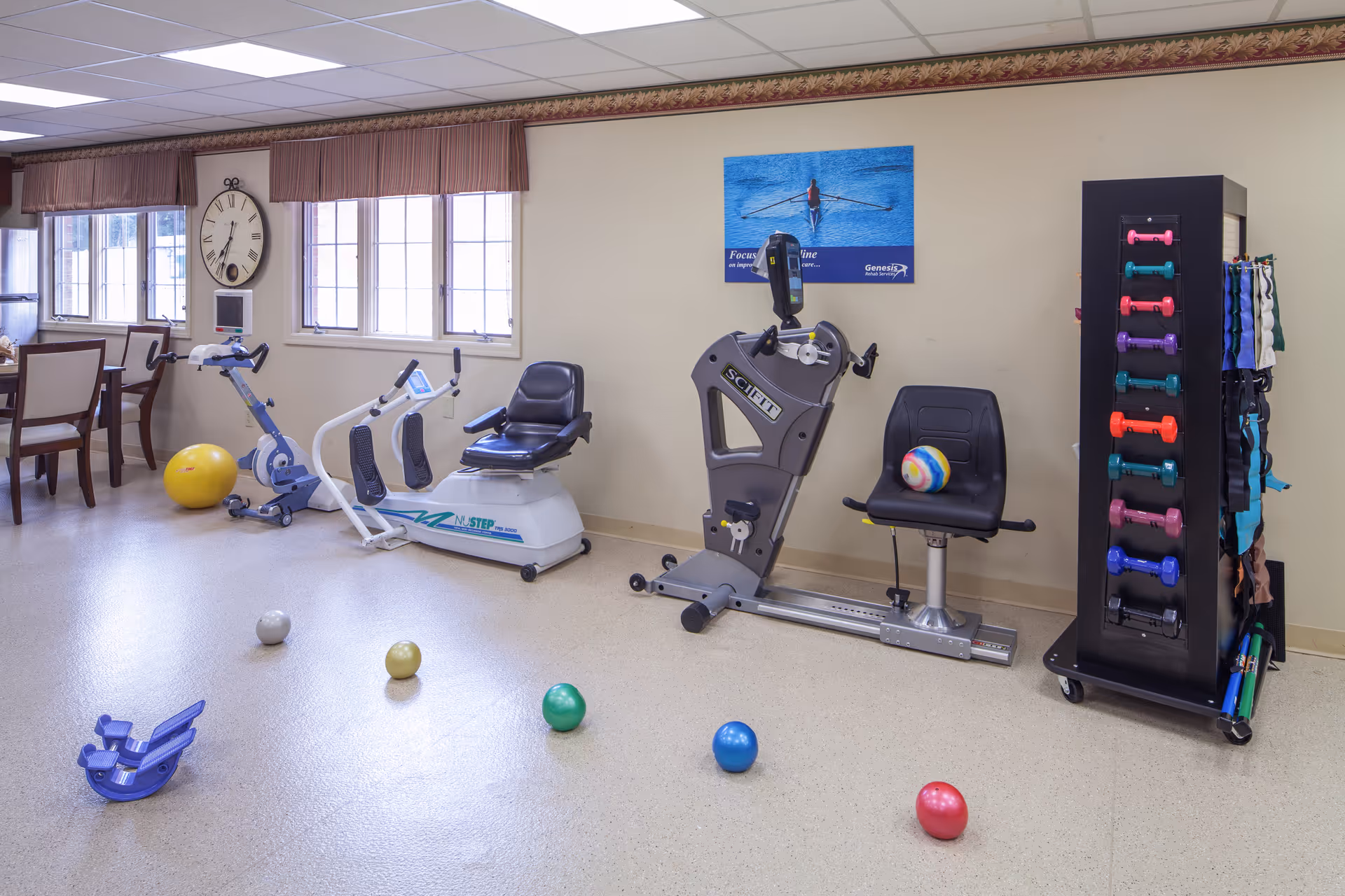 A fitness room in a senior living facility with exercise equipment including stationary bikes, a rowing machine, colorful dumbbells on a rack, and various exercise balls on the floor. There is a large clock on the wall and windows letting in natural light. A motivational poster is displayed on the wall above the equipment.