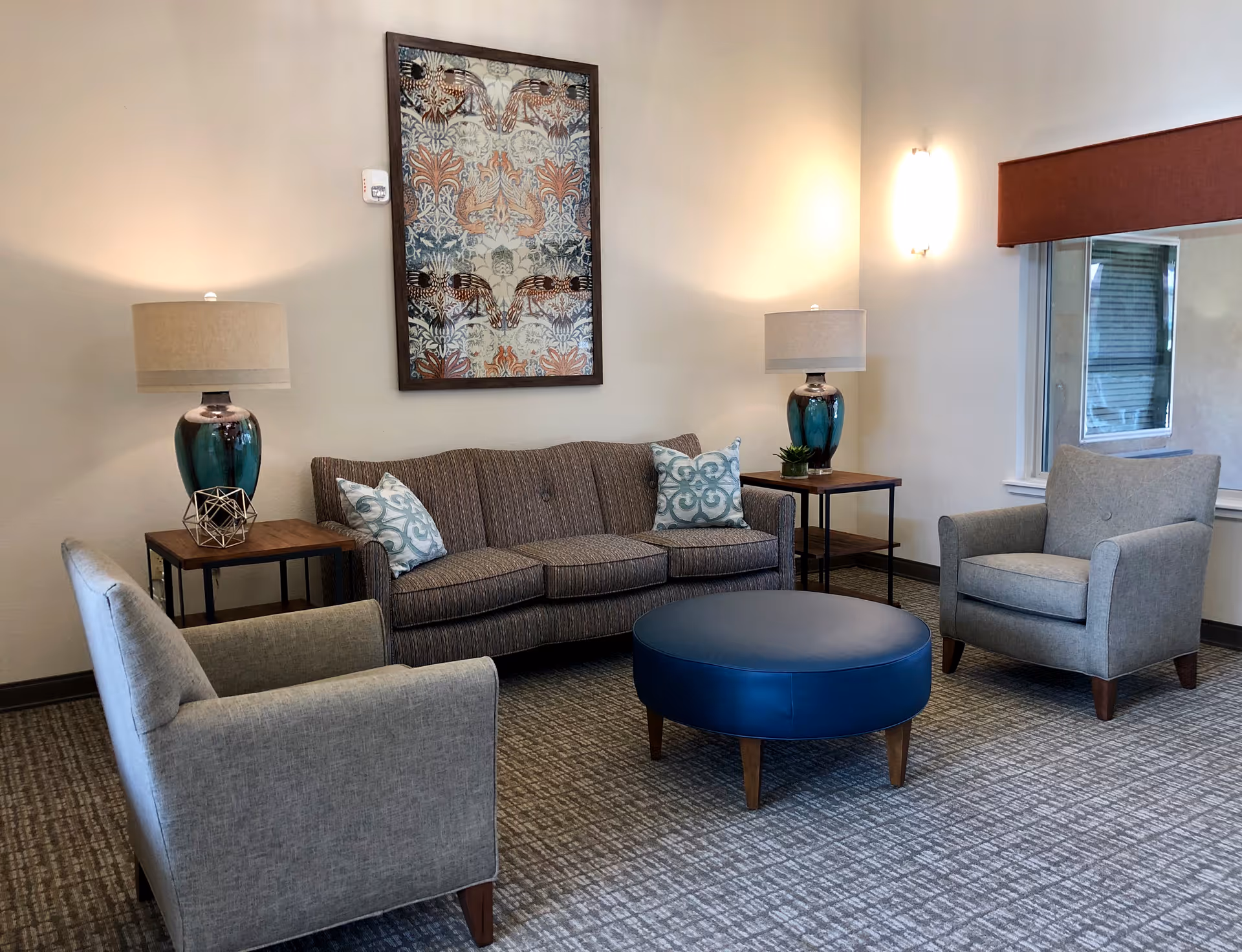 A cozy living room area featuring a brown striped sofa with two patterned throw pillows, two gray armchairs, a round blue ottoman, two side tables each with a teal lamp, and a framed decorative artwork on the wall. There is a window with a brown valance on the right side.