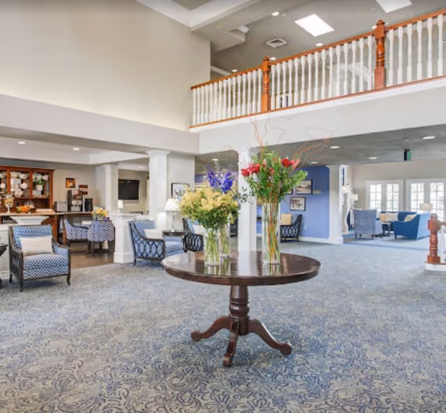 Spacious and well-lit senior living facility common area with a round wooden table in the center holding three vases of colorful flowers. The room features patterned carpet, comfortable armchairs with cushions, a wooden railing on the upper level, and a seating area with blue and white furniture in the background.