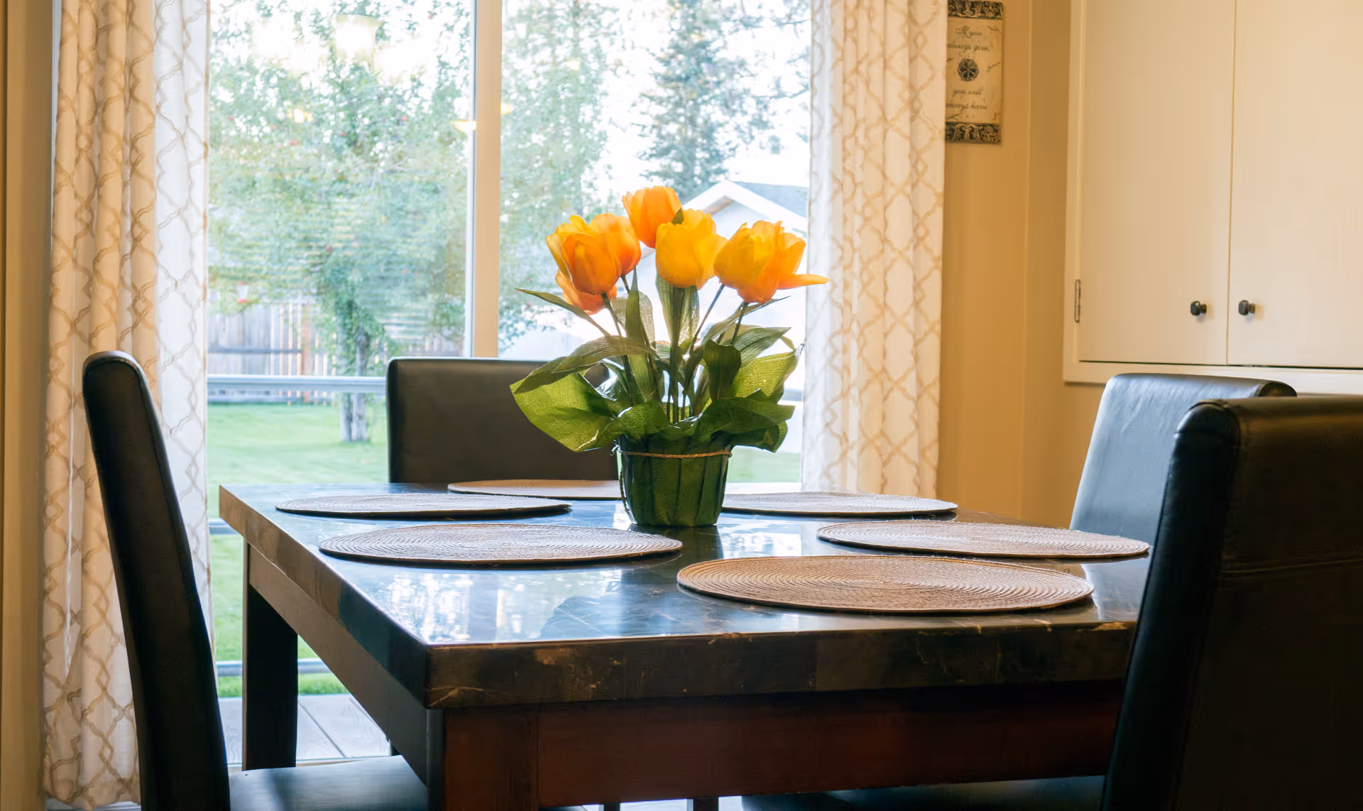 A dining table with four black chairs around it, set with four round woven placemats and a vase of yellow and orange tulips in the center. Behind the table is a large window with patterned curtains, showing a green outdoor yard with trees and a fence.