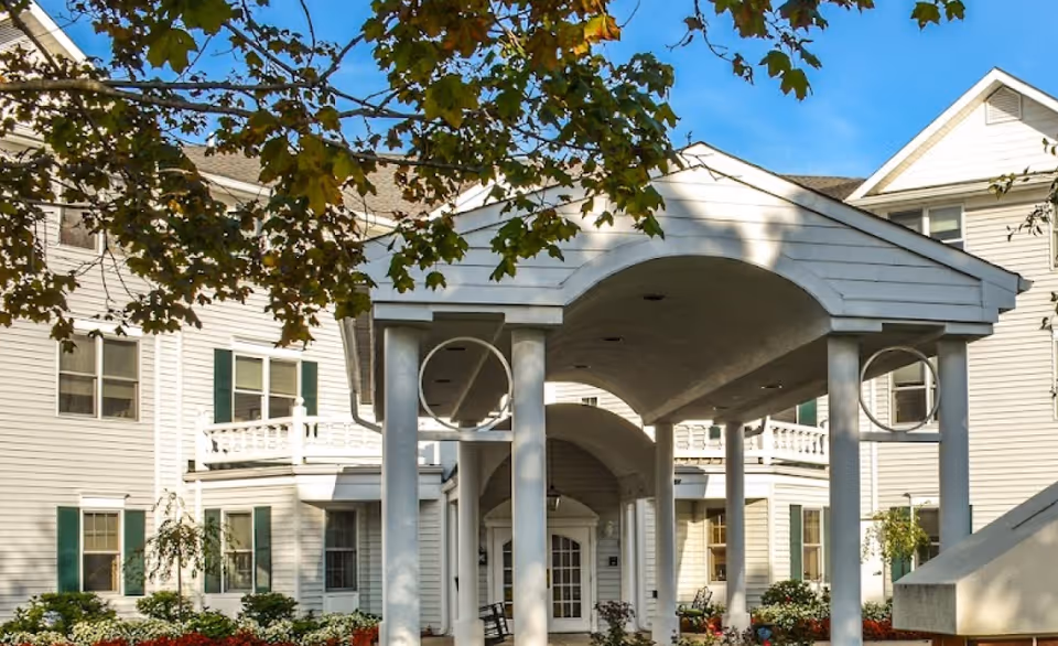 Exterior view of a white multi-story assisted living facility with green shutters, a covered entrance supported by white columns, and landscaping with flowers and trees under a blue sky.