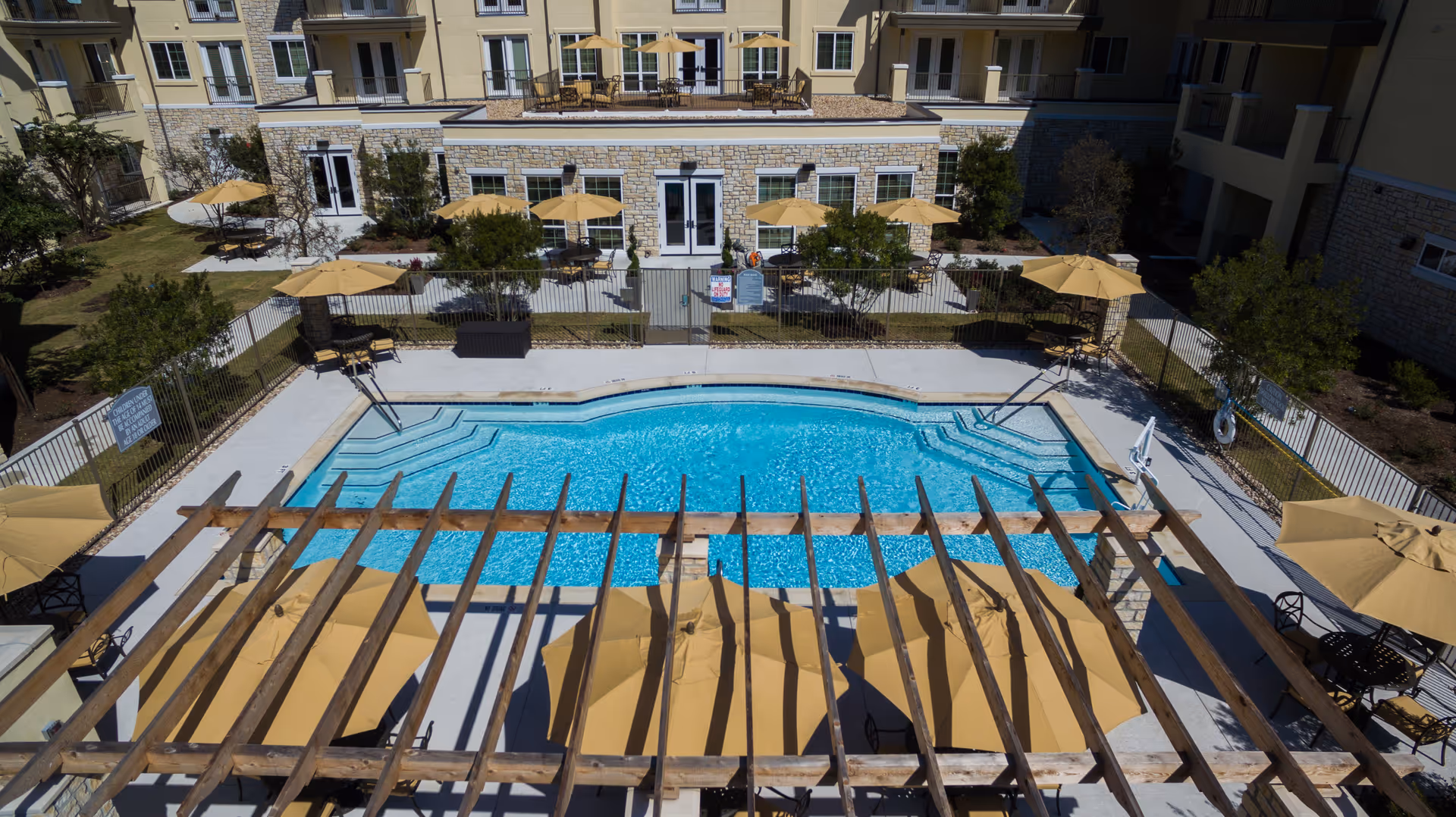 Aerial view of a swimming pool area at Lake Travis Independent Living facility, surrounded by a fence and several yellow umbrellas with tables and chairs. The pool area is adjacent to a multi-story building with stone and beige exterior walls, balconies, and patio seating.