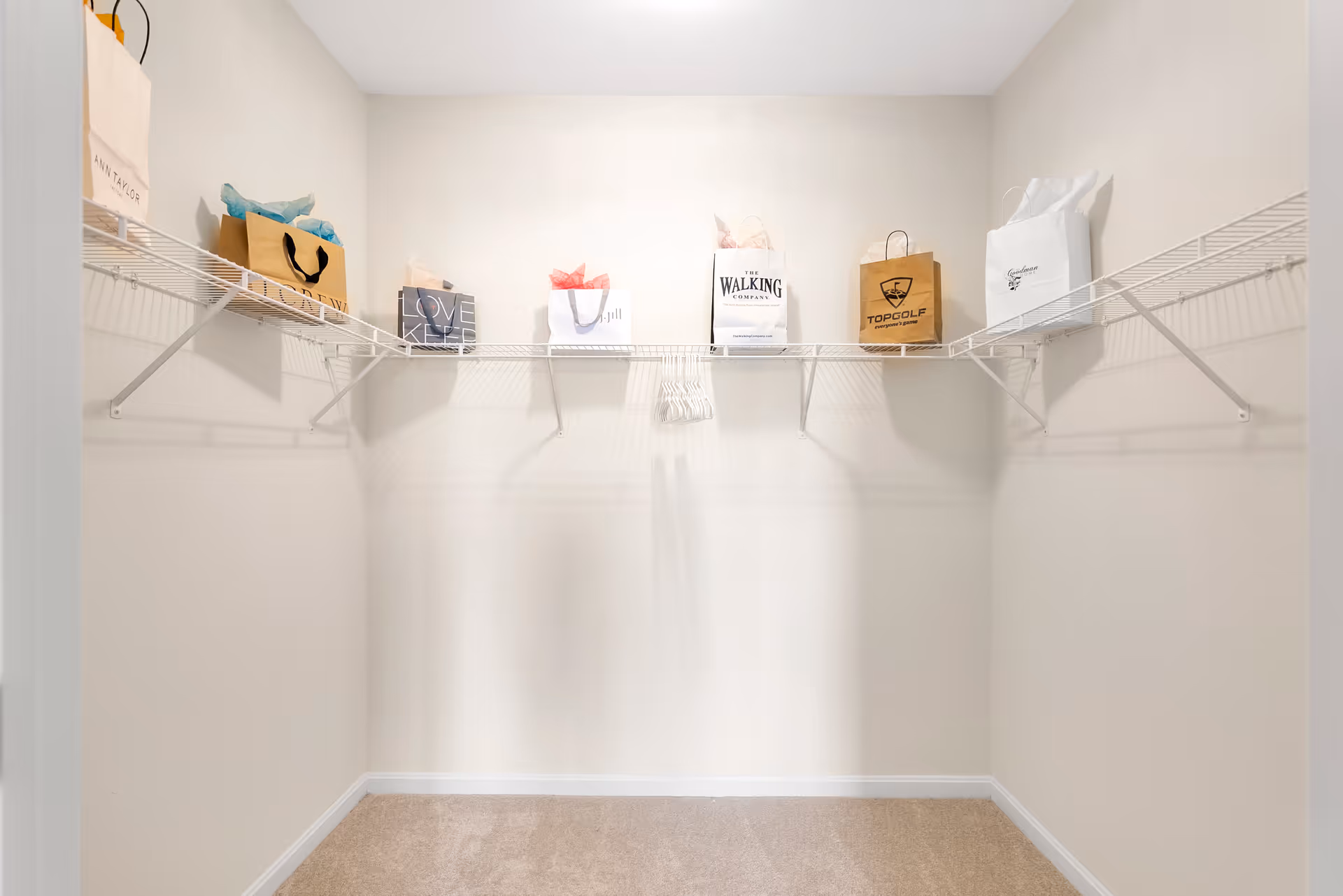 Empty walk-in closet with white wire shelving on three walls. Several shopping bags are placed on the shelves, and a few white hangers are hanging in the center. The closet has beige carpet flooring and light-colored walls.