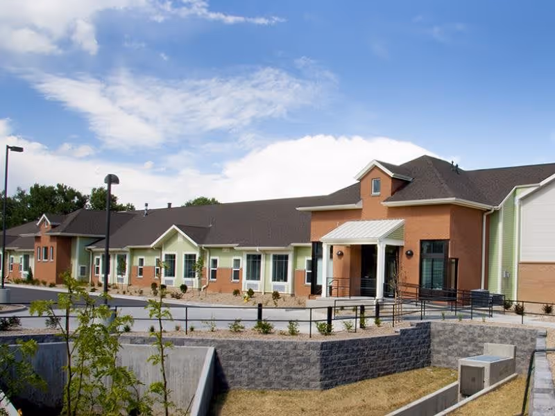 Exterior view of a single-story assisted living facility building with a combination of brick and light green siding, multiple windows, and a covered entrance. The sky is partly cloudy and there are some small plants and landscaping in front of the building.