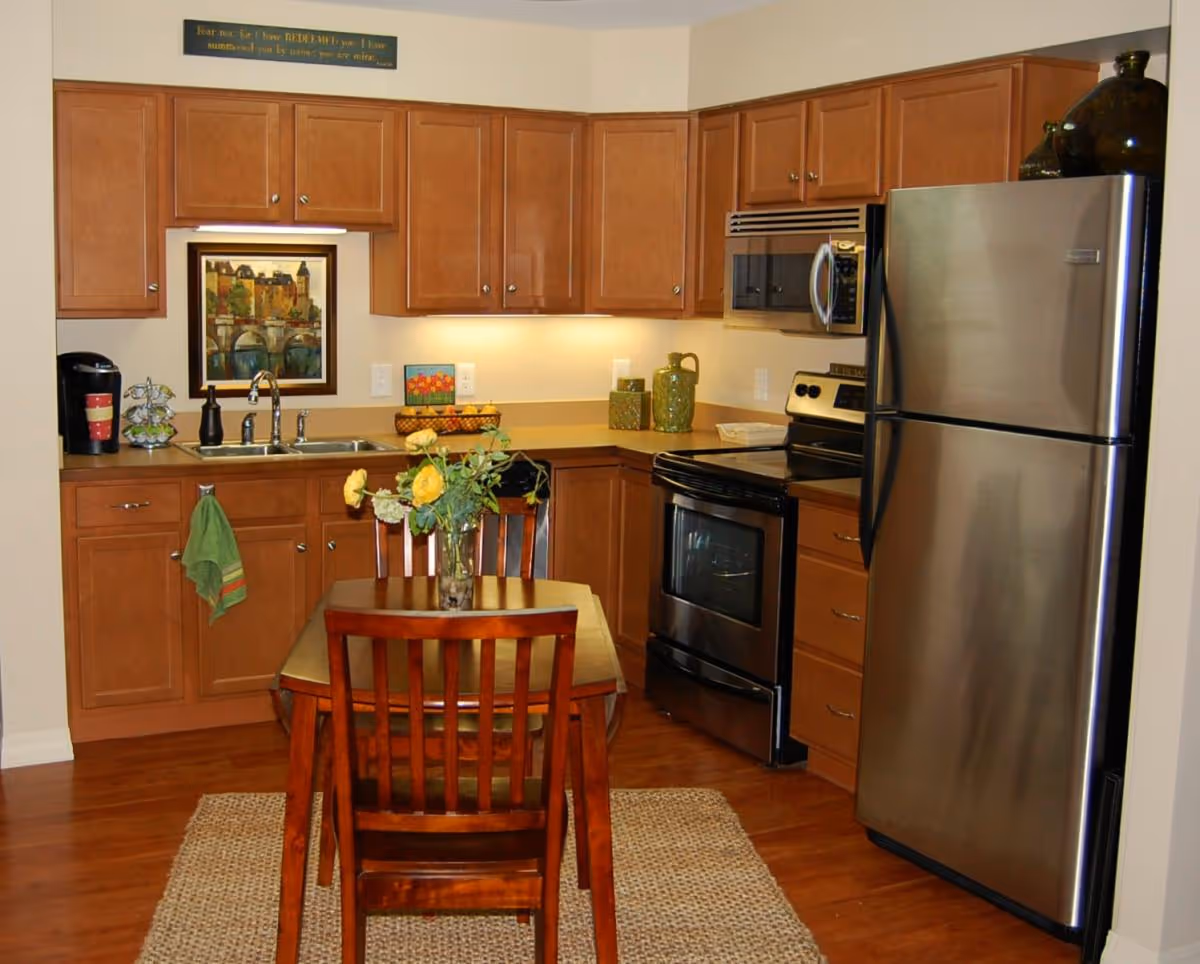 Cozy kitchen with wooden cabinets, stainless steel refrigerator and stove, and a small wooden dining table with a vase of yellow flowers.