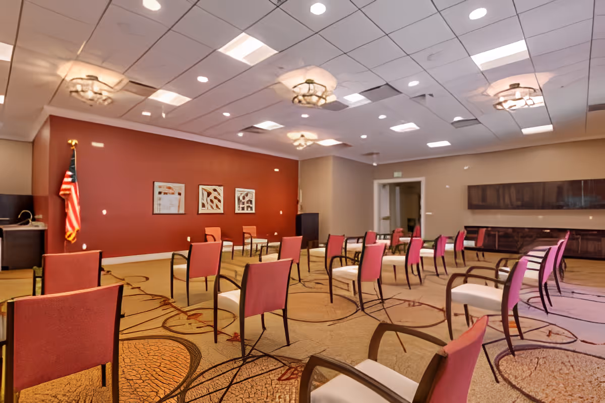 A spacious meeting or activity room with multiple chairs arranged in rows facing a red accent wall. The room features a patterned carpet, ceiling lights, an American flag in the corner, and framed artwork on the wall.