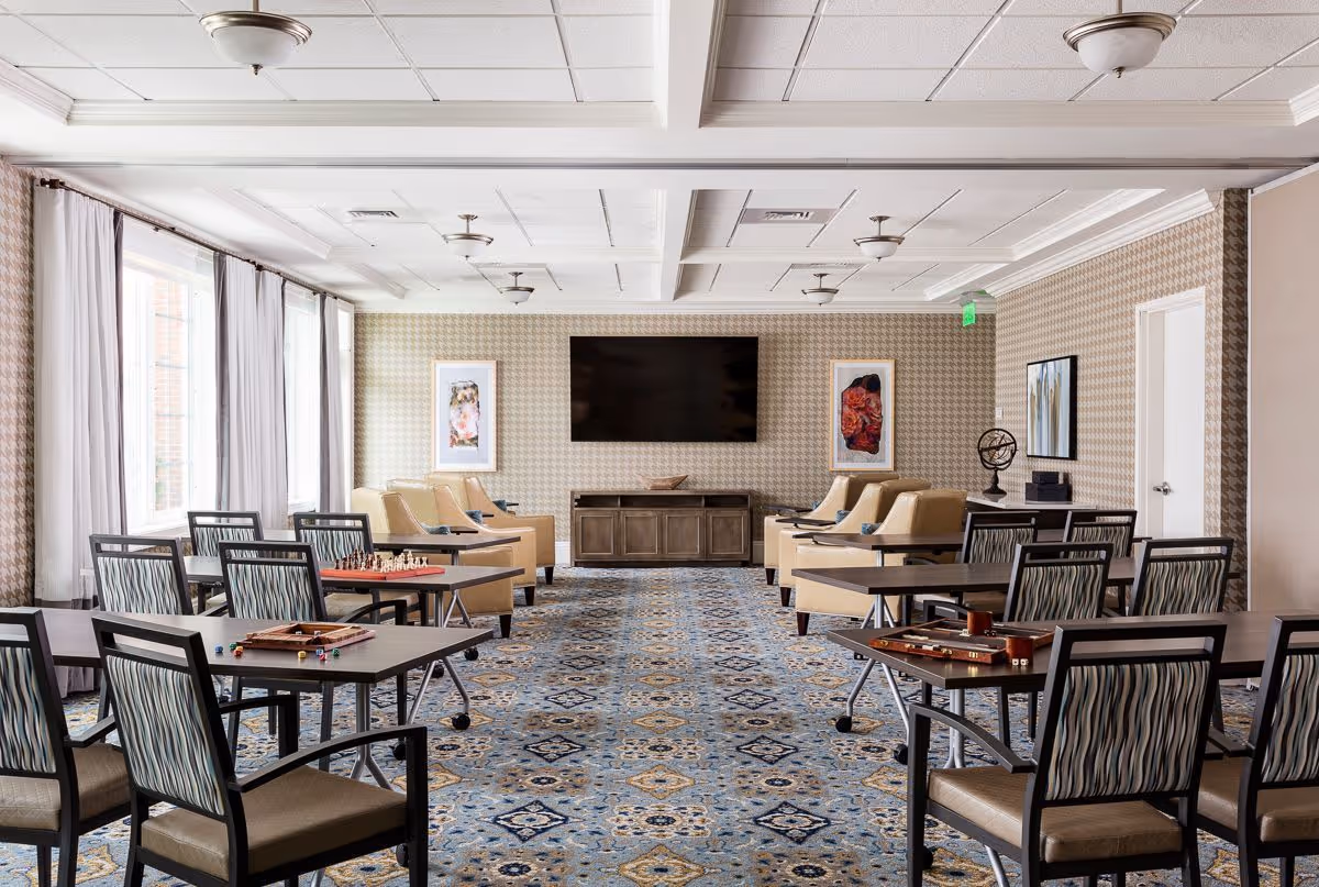 A spacious and well-lit common room with multiple tables and chairs arranged for games and social activities. The room features a patterned carpet, beige armchairs along the back wall, a large flat-screen TV mounted on the wall, and framed artwork on either side of the TV. Large windows with white curtains allow natural light to fill the space.