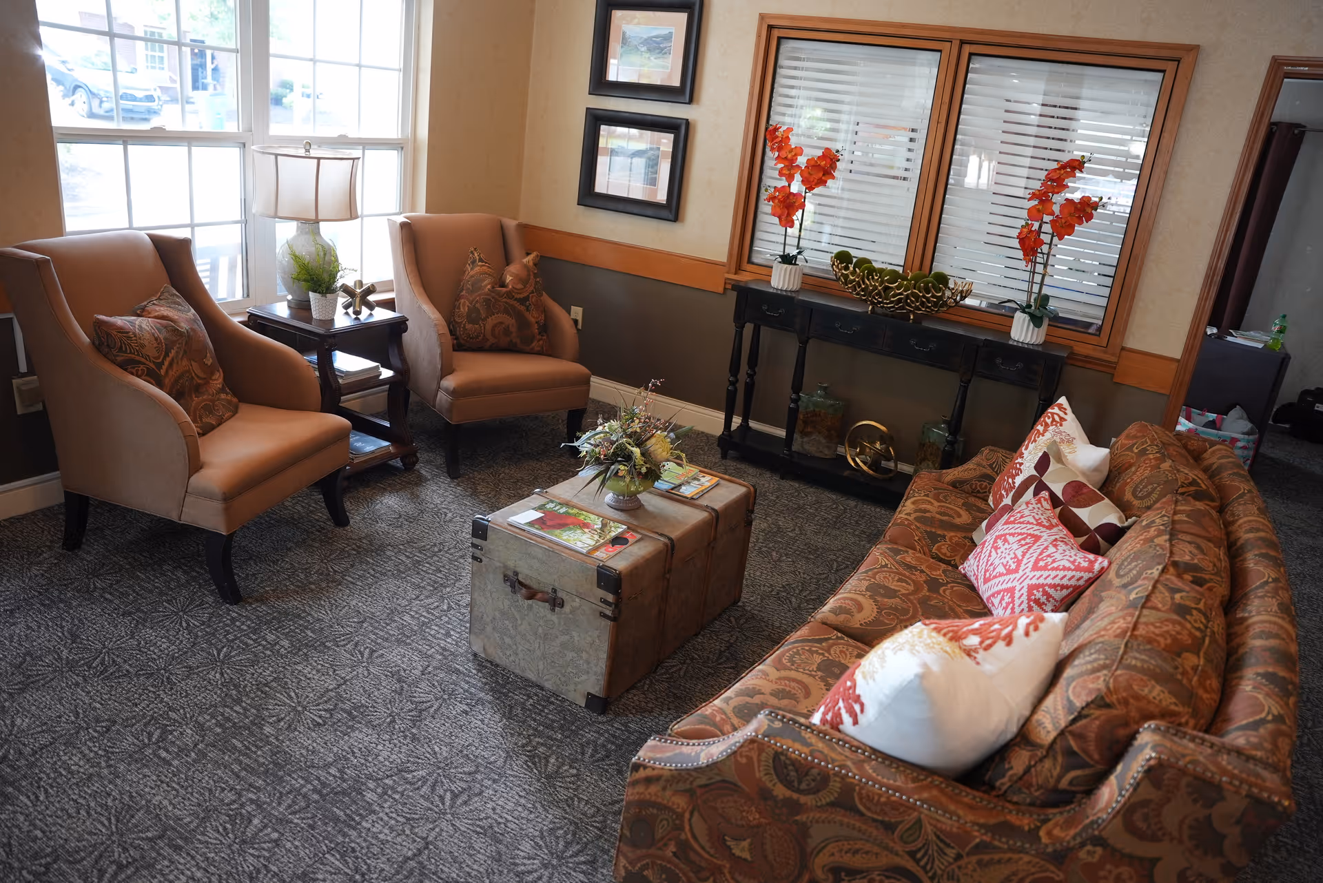 A cozy living room area with a patterned sofa adorned with decorative pillows, two beige armchairs with patterned cushions, a vintage trunk used as a coffee table with magazines and a floral arrangement on top, a side table with a lamp and plant, and a black console table against the wall with two vases of orange flowers and decorative items. Large windows with blinds and framed pictures on the wall complete the room.