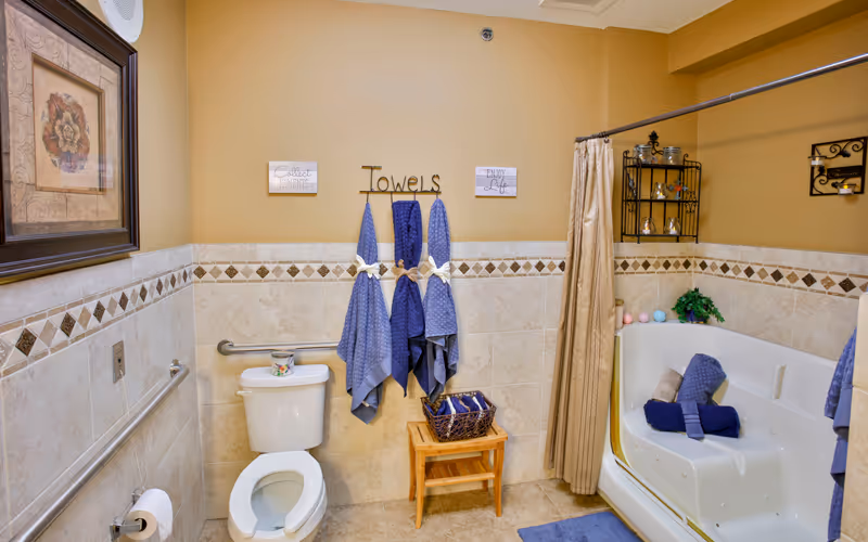 Bathroom with a toilet, a tub/shower, blue towels hanging on hooks labeled "Towels", and beige tiled walls and floor.