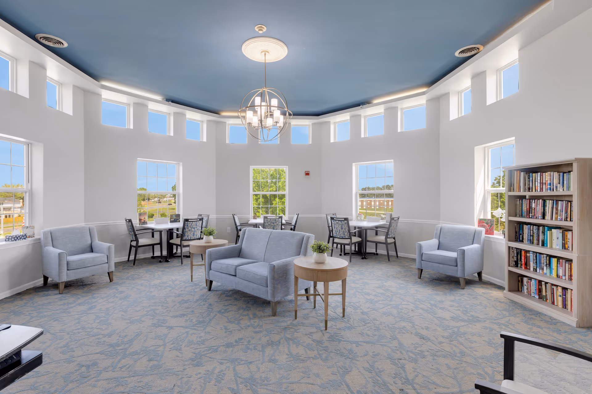 A bright and spacious common area with large windows letting in natural light. The room features a blue ceiling with a modern chandelier, light gray armchairs and a loveseat, small round wooden tables with potted plants, and several tables with chairs near the windows. A bookshelf filled with books is visible on the right side.