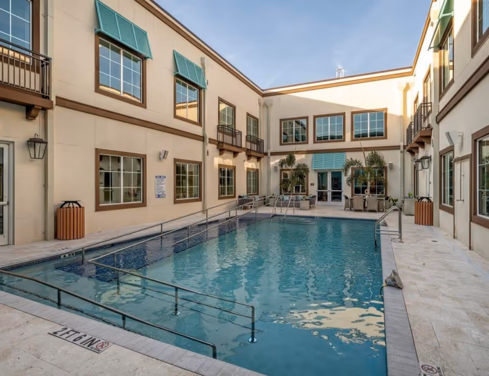 Outdoor swimming pool area at Sunscape Boca Raton facility, surrounded by a two-story building with multiple windows and green awnings. The pool has handrails and a ramp for accessibility, with seating and potted plants near the entrance doors.