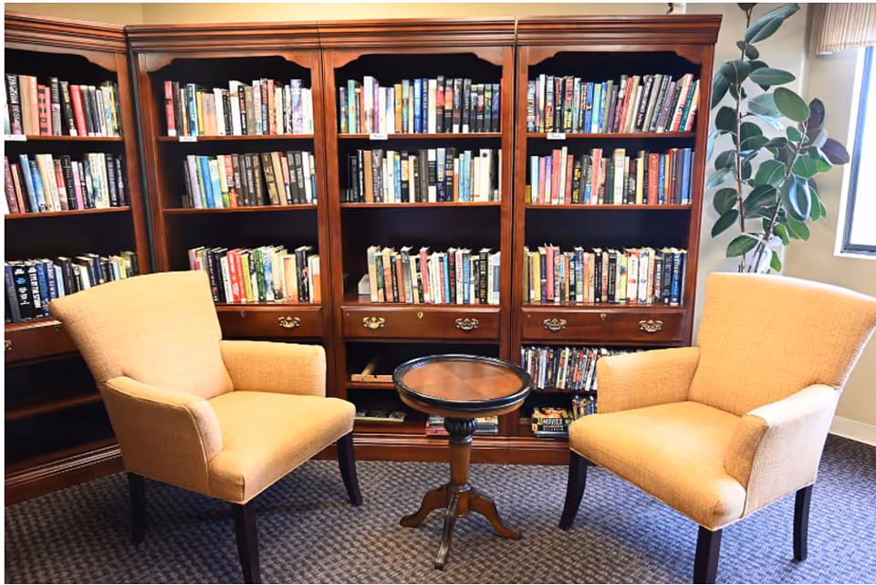 A cozy reading area with two beige upholstered armchairs facing each other with a small round wooden table between them. Behind the chairs is a large wooden bookshelf filled with various books. A potted plant is visible near a window on the right side.