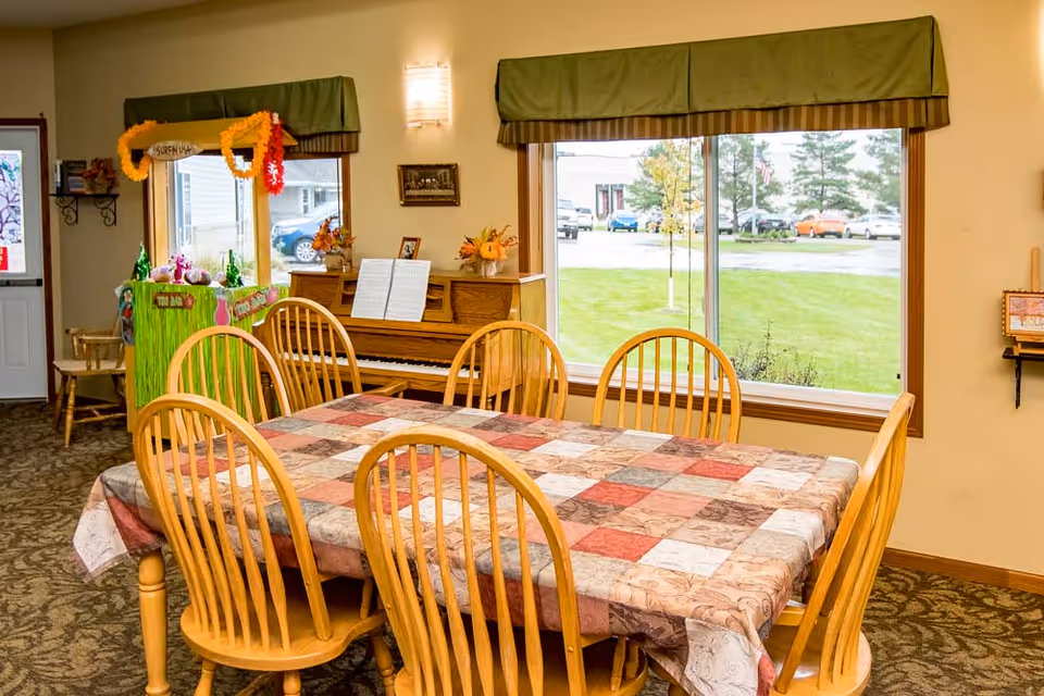 A dining area with a wooden table covered by a patchwork tablecloth and six wooden chairs. Behind the table, there is an upright piano with sheet music and flower decorations on top. To the left, there is a small colorful stand decorated with leis and a 'SURF N' sign. Large windows with green valances show a grassy outdoor area and parked cars outside.