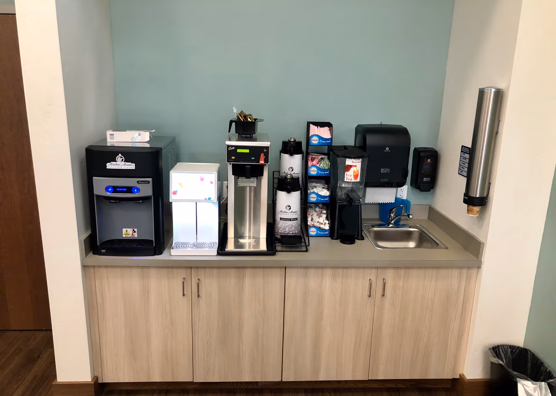 A beverage station with a coffee machine, ice dispenser, hot water dispenser, cups, stirrers, napkins, a small sink, and a soap dispenser mounted on the wall. The station is set against a light blue wall with light wood cabinets below the countertop.