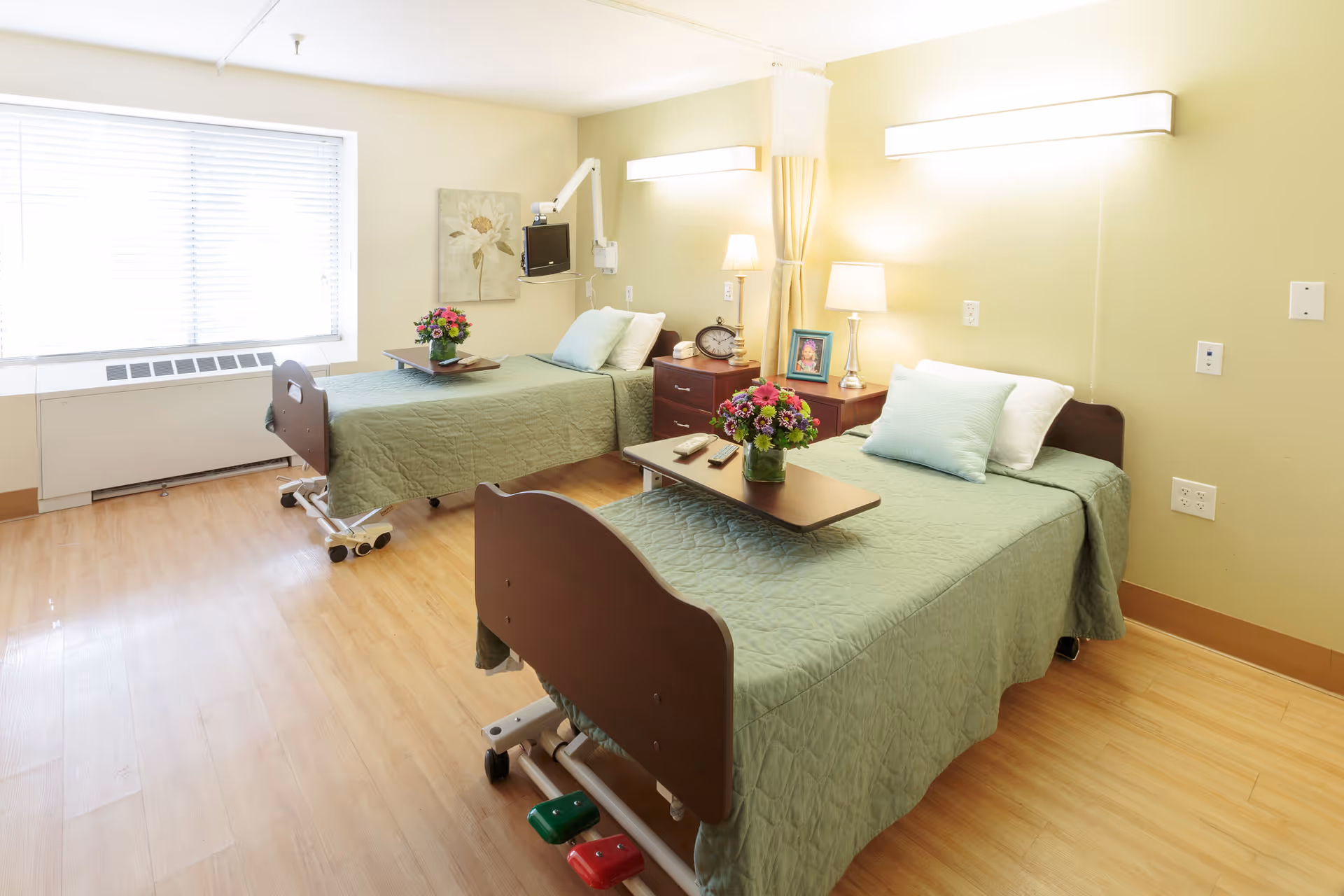 A bright and clean rehabilitation center room with two hospital-style beds covered in green quilts, each with a pillow and a small table holding a vase of flowers. The room has light-colored walls, wooden flooring, a large window with blinds, bedside tables with lamps, a clock, and a framed photo. Medical equipment is mounted on the wall near one bed.