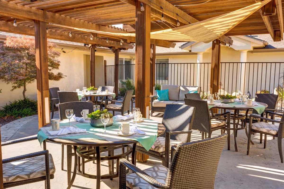 Outdoor patio area with wooden pergola providing partial shade over several round tables set for dining. The tables have green tablecloths, white napkins, cups, and glassware. Wicker chairs with cushions surround the tables, and there is a cushioned seating area with turquoise pillows in the background. The space is enclosed by a black metal fence and has some landscaping with plants and a tree nearby.