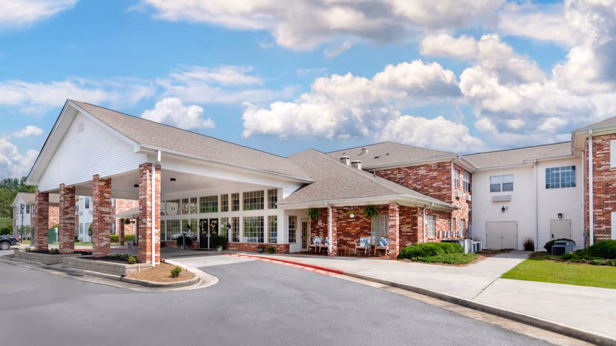 Exterior view of a senior living facility building with a covered entrance supported by brick columns, large windows, and a driveway in front. The sky is partly cloudy.