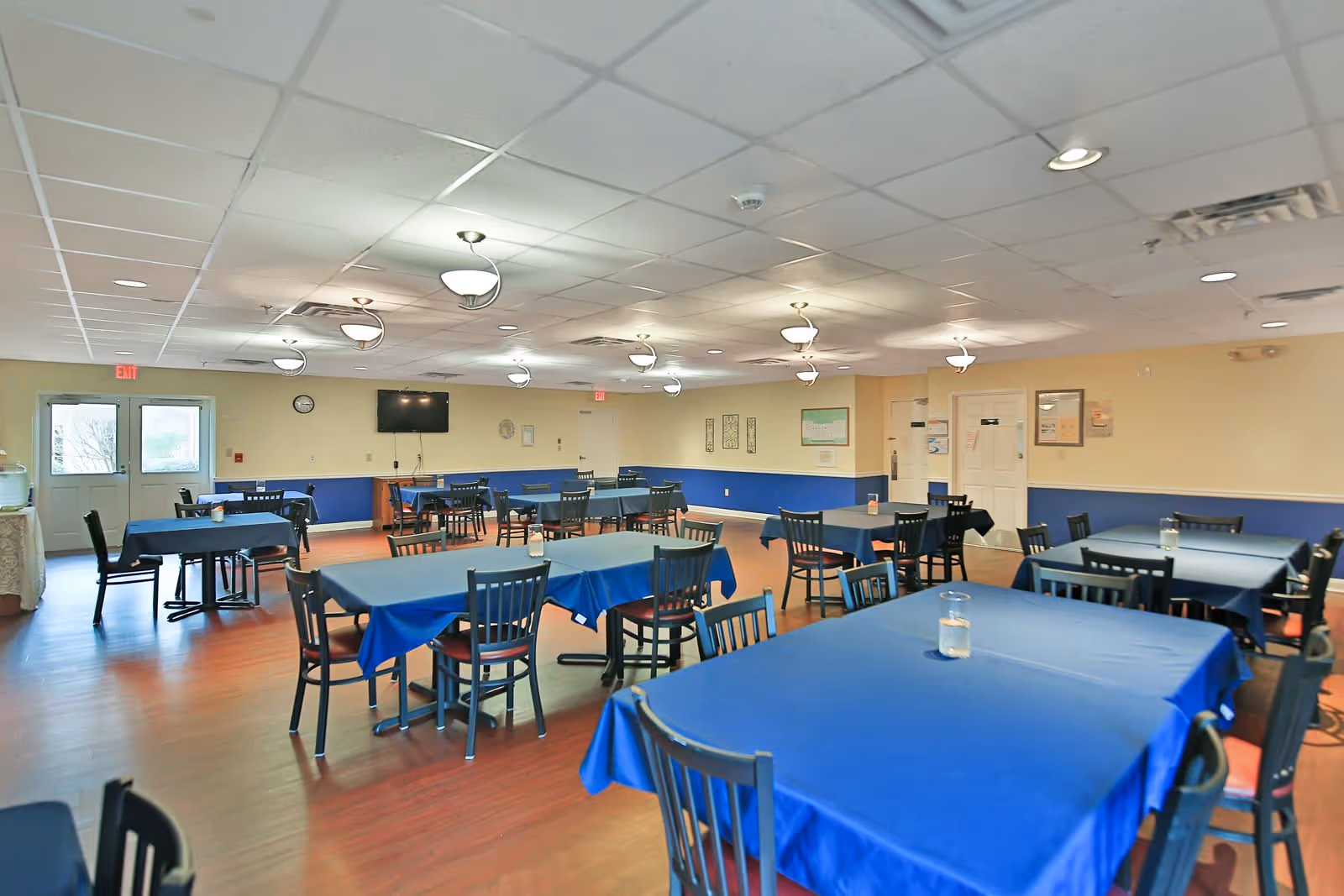 A spacious dining room with multiple tables covered in blue tablecloths and black chairs arranged around them. The room has a wooden floor, beige walls with a blue lower half, and a white tiled ceiling with modern light fixtures. There is a wall-mounted TV and a clock on the far wall, and double doors with exit signs above them.