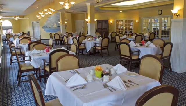 A spacious dining room with multiple tables covered in white tablecloths, each set with napkins, cups, and silverware. The room features beige walls, carpeted floors with a patterned design, and several chairs with cushioned seats and backs. Soft lighting fixtures hang from the ceiling and walls, and there are large windows and glass doors along one side of the room.