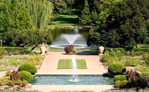 Formal landscaped garden with tiered reflecting pools and fountains, urns, manicured bushes, and trees leading to a distant pond.