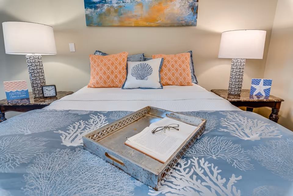 A neatly made bed with a blue and white coral-patterned bedspread and three decorative pillows, two orange and one white with a shell design. On the bed is a metal tray holding an open book and a pair of reading glasses. On each side of the bed are wooden nightstands with matching lamps, a small clock, and decorative items including a sign that reads 'Happiness comes in waves' and a starfish-themed picture. A colorful abstract painting hangs on the wall above the bed.