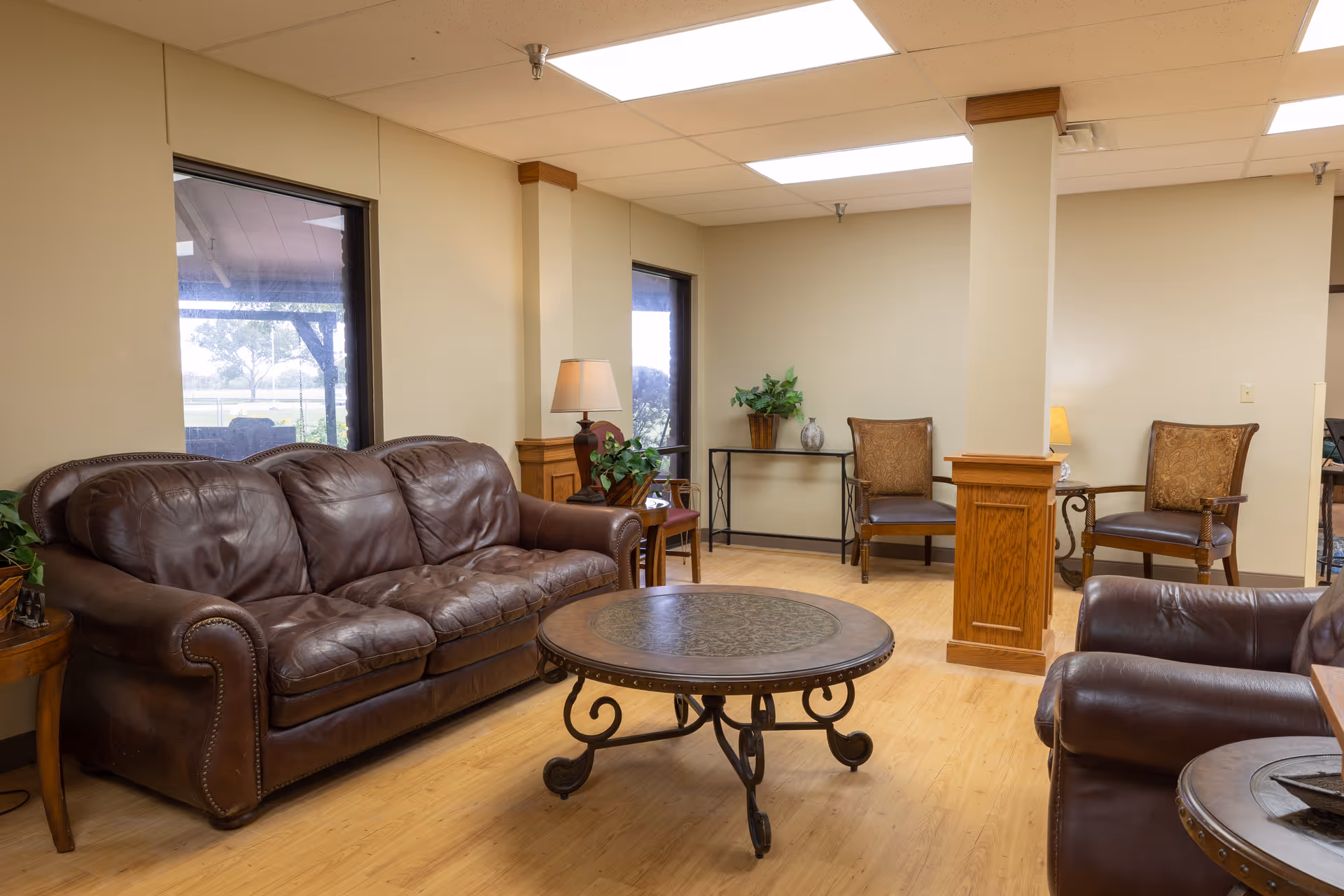 A cozy living room area in a senior living facility featuring two brown leather sofas, a round metal and wood coffee table, two wooden armchairs with cushions, wooden side tables with lamps, and potted plants. Large windows provide natural light and a view of the outdoors.