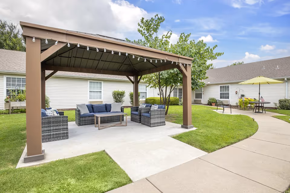 Outdoor seating area with a brown wooden pergola covering a set of wicker furniture including a sofa and chairs with cushions, situated on a concrete slab surrounded by green grass and a curved sidewalk. In the background, there are single-story white buildings with windows and some outdoor furniture including a table with a yellow umbrella.