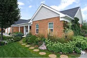 Brick building entrance with a pitched roof, landscaped flower beds and circular stepping stones leading to the doorway under a blue sky.