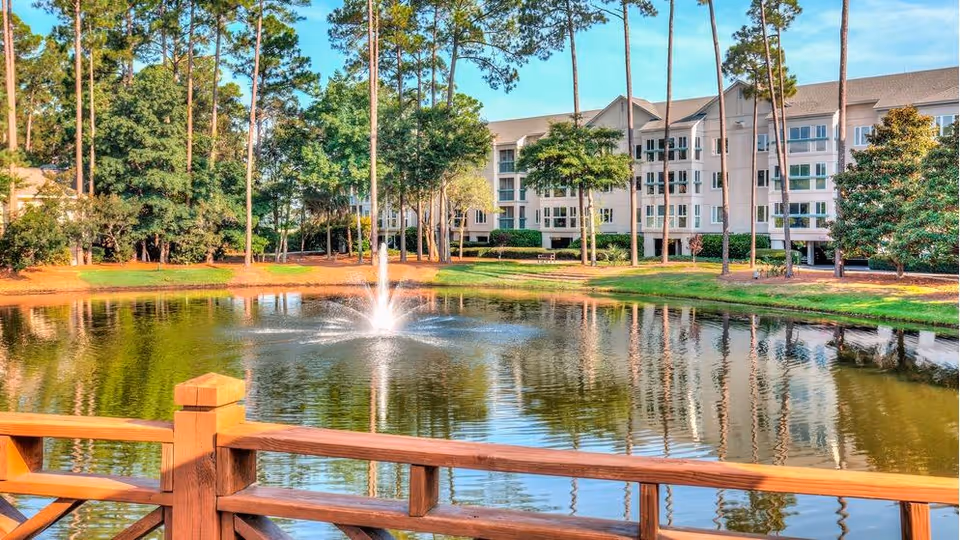 Pond with a central fountain and wooden railing in the foreground, with a multi-story residential building and tall trees in the background.