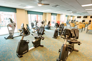A spacious fitness room with several stationary exercise bikes and other cardio equipment arranged on a carpeted floor. Large windows along one wall allow natural light to fill the room. In the background, there are tables and chairs, suggesting a multi-use space.